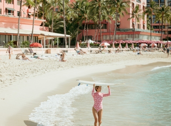 A woman carrying a surfboard walks along the beach in front of The Royal Hawaiian.