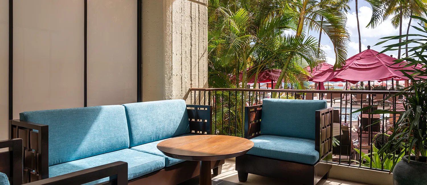 Balcony furnished with blue sofas and a wooden table, overlooking the pool area with pink umbrellas.