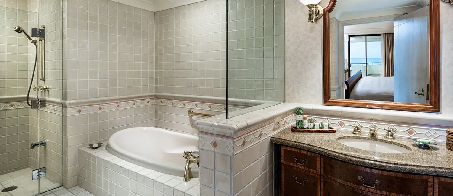 The Ali‘i Suite bathroom with a bathtub and vanity, showing the bedroom and ocean view in the mirror.