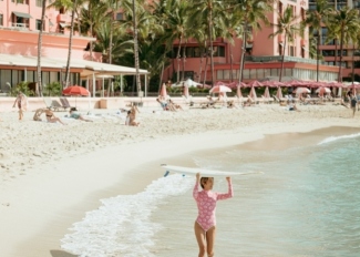 A woman carrying a surfboard walks along the beach in front of The Royal Hawaiian.